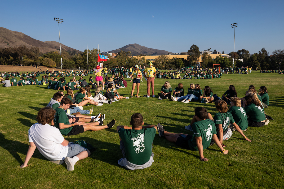 A large group of students sits in a circle on a field as their WOW leaders stand and address them