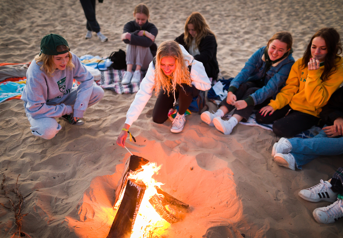 Students in Cal Poly sweatshirts roast marshmallows over a bonfire on the beach