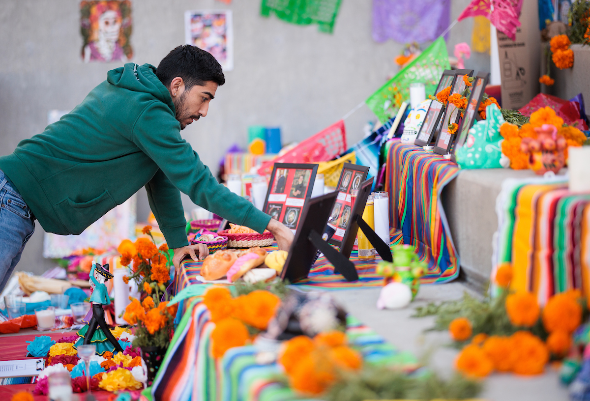 A young man in a green sweatshirt arranges items on a brightly-decorated ofrenda at the University Union plaza