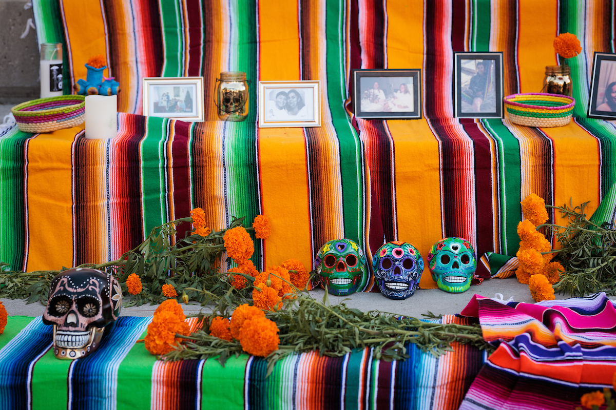 A close-up shot of an ofrenda, draped with bright striped cloth and featuring painted skulls, vintage photos and bouquets of marigolds