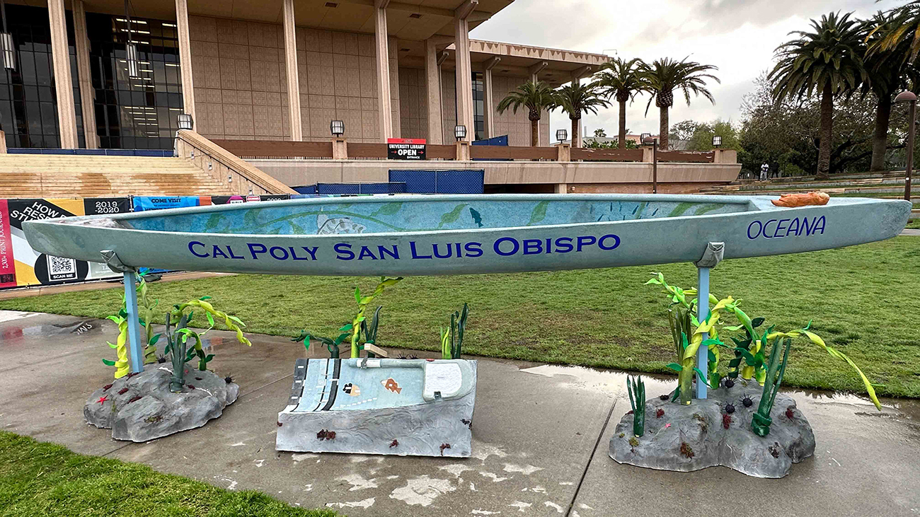 A long blue canoe painted with the words "Cal Poly San Luis Obispo" and "Oceana" rests on mounts decorated with fake seaweed in front of a university building.