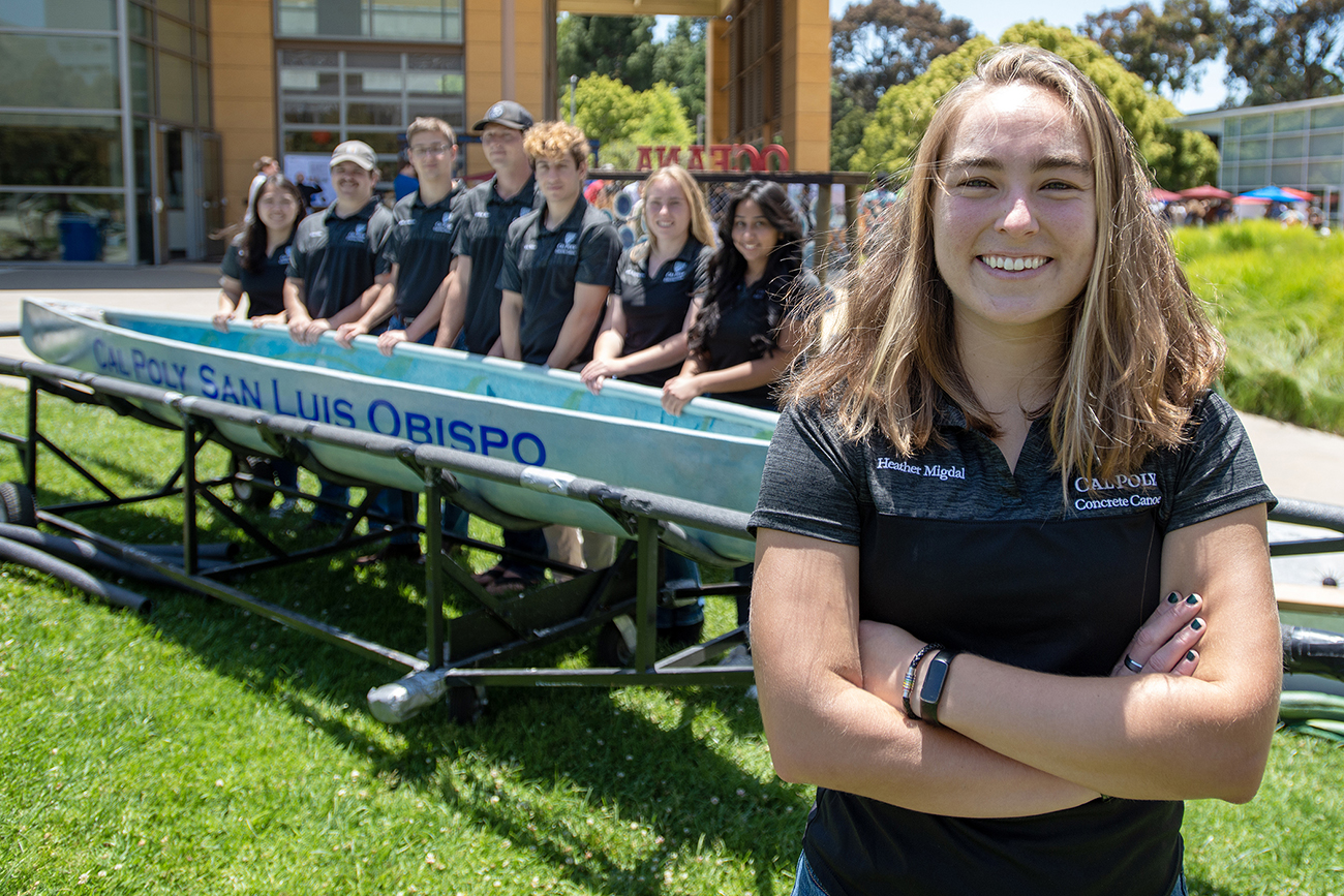 A student in a black team polo shirt poses with arms crossed in front of a canoe and her teammates