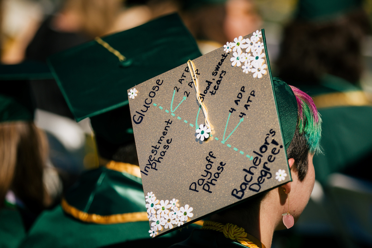 A student with colorful hair sports a mortarboard cap with a yellow tassel decorated with flowers and a faux-scientific formula celebrating the path to graduation