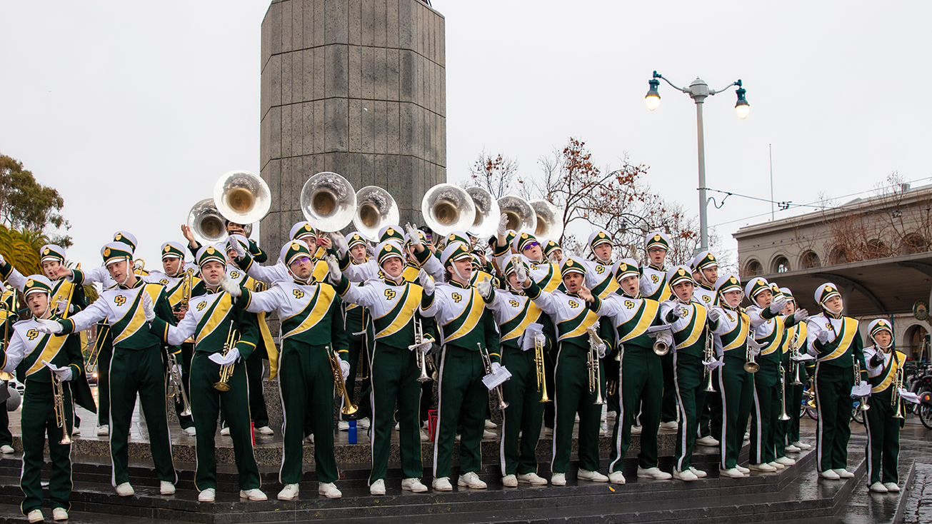 Members of the Mustang Band, in white, yellow and green uniforms, perform in front of a concrete column at San Francisco's Embarcadero 
