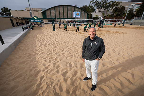 A man in a Cal Poly windbreaker stands in front of a sand-covered beach volleyball court while young women in tracksuits practice in the background
