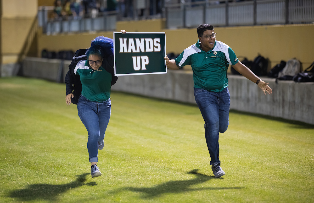 Two student leaders run across the field at Spanos Stadium holding a sign reading Hands Up