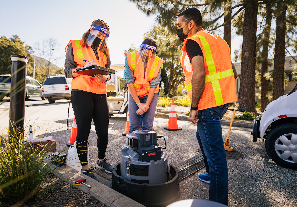 Three people in masks and orange safety vests work on a robotic device next to a manhole cover
