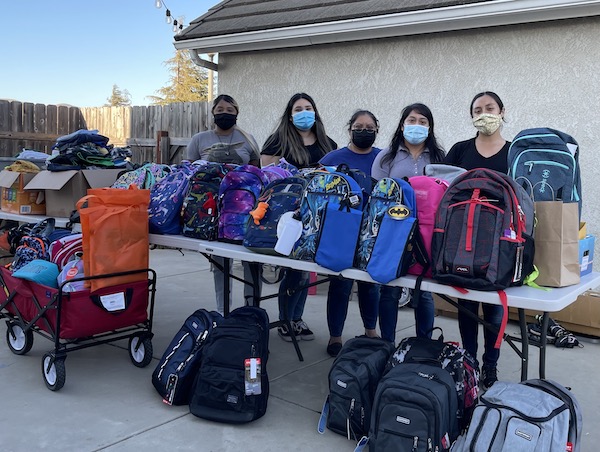 Masked students stand behind backpacks filled with school supplies for farmworkers' families.
