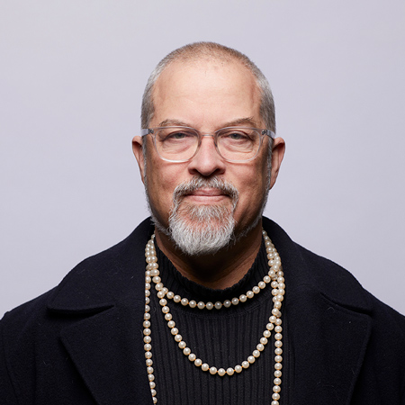 A person with a beard wears glasses, a black coat and a string of pearls