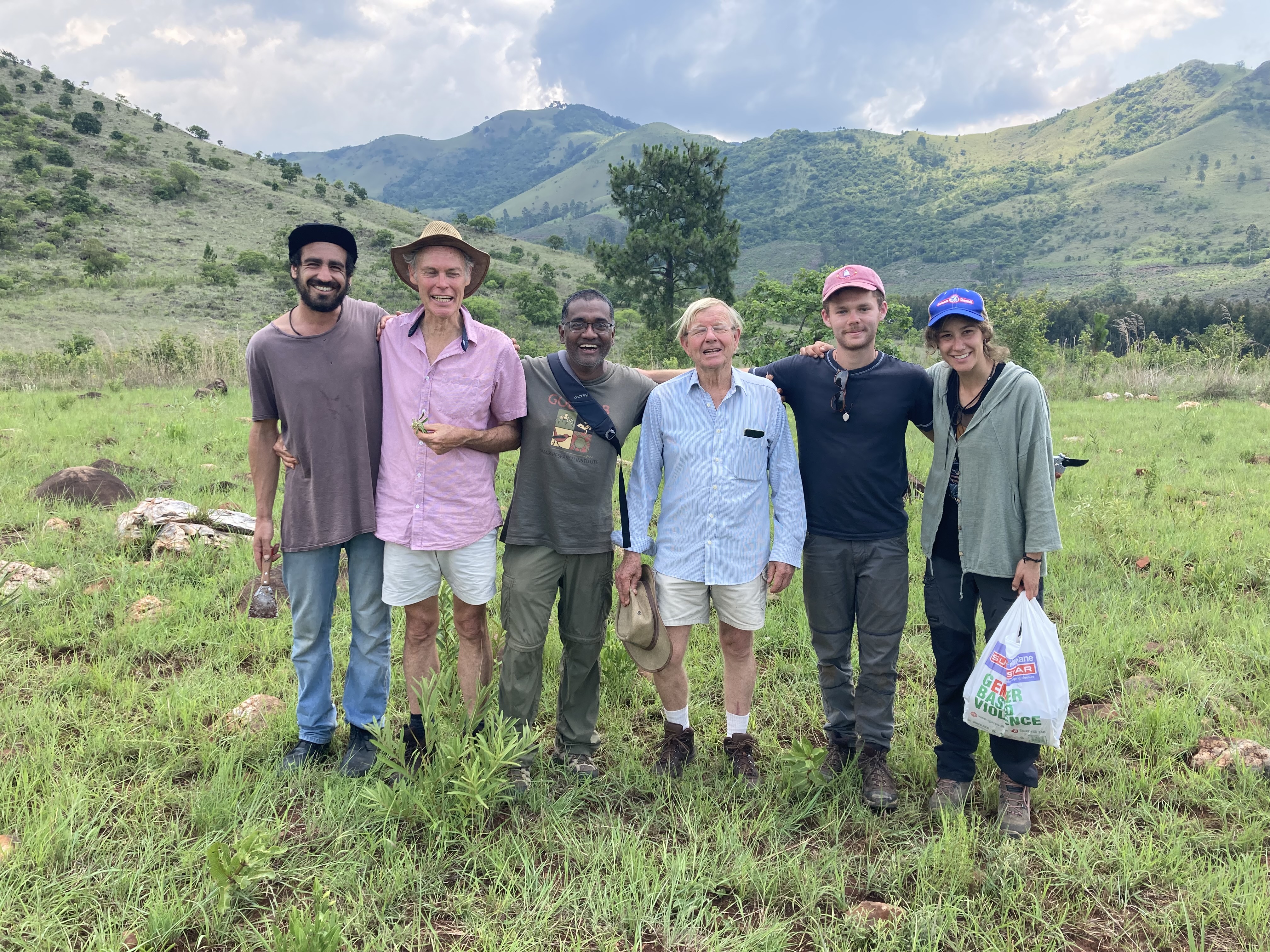 Professor Nishi Rajakaruna and student TJ Samojedny posing with a group of colleagues in a field surrounded by mountains in South Africa.
