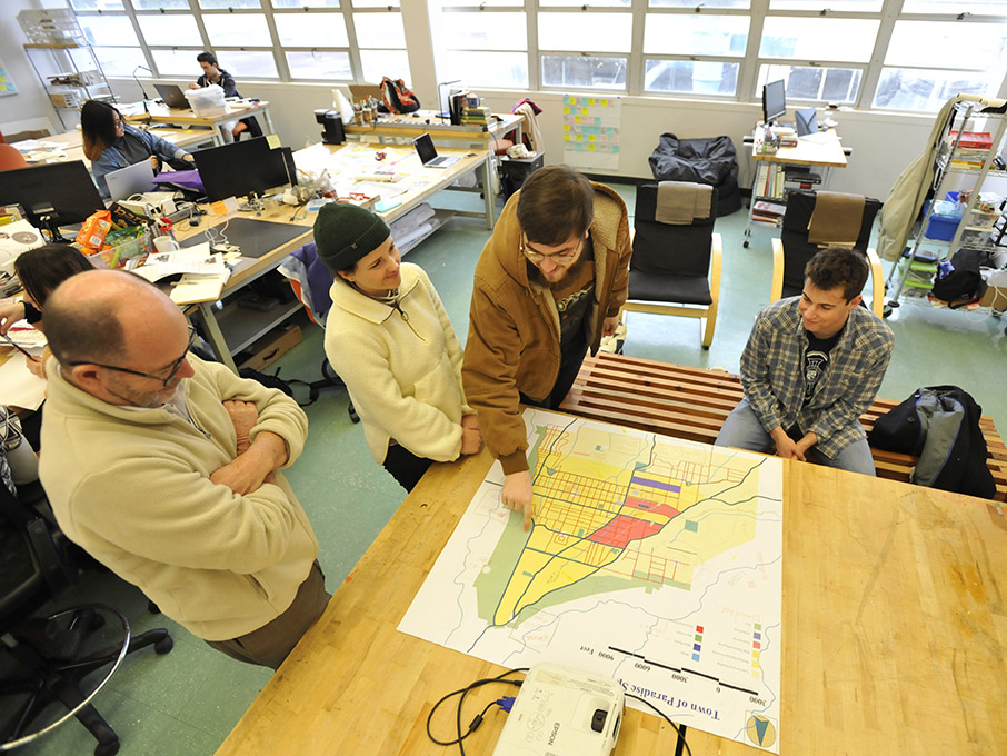 Four people stand around a table in a classroom while one of them points to a spot on a map lying on the table. 