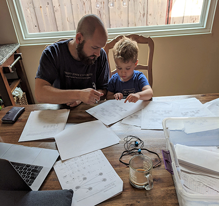 Professor Paul Anderson, sitting with his young son, grades papers at his dining room table.