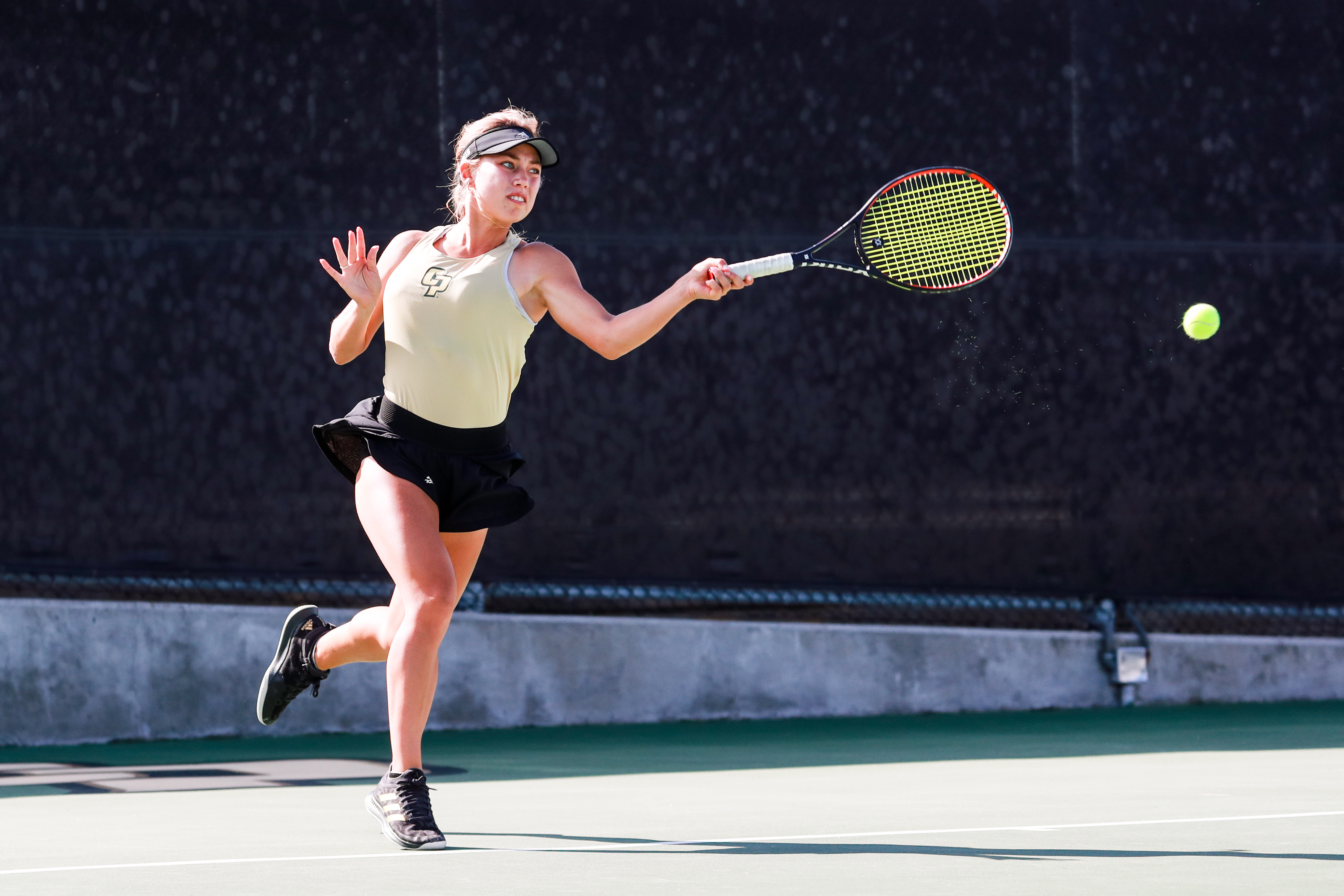 A blonde woman wearing a Cal Poly tennis uniform hits a ball with her racket on the court. 