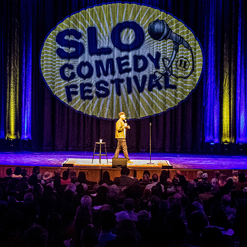 A man stands on a stage with a large yellow and blue projection that reads “SLO Comedy Festival” with a crowd of people in front of him.