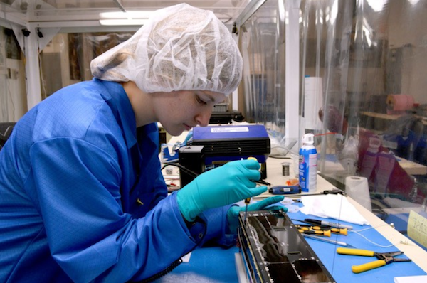 A woman in a blue coat, teal latex gloves and a white scrub cap uses a screwdriver to work on the ExoCube 2 CubeSat.