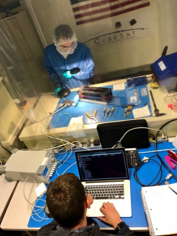 A man in a blue coat, white scrub cap and teal gloves tests the ExoCube 2 satellite while on the other side of a clear partition a man looks at a laptop.