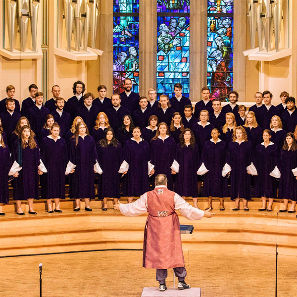 The choir of over 40 people in navy robes, stands in a semi-circle facing the conductor