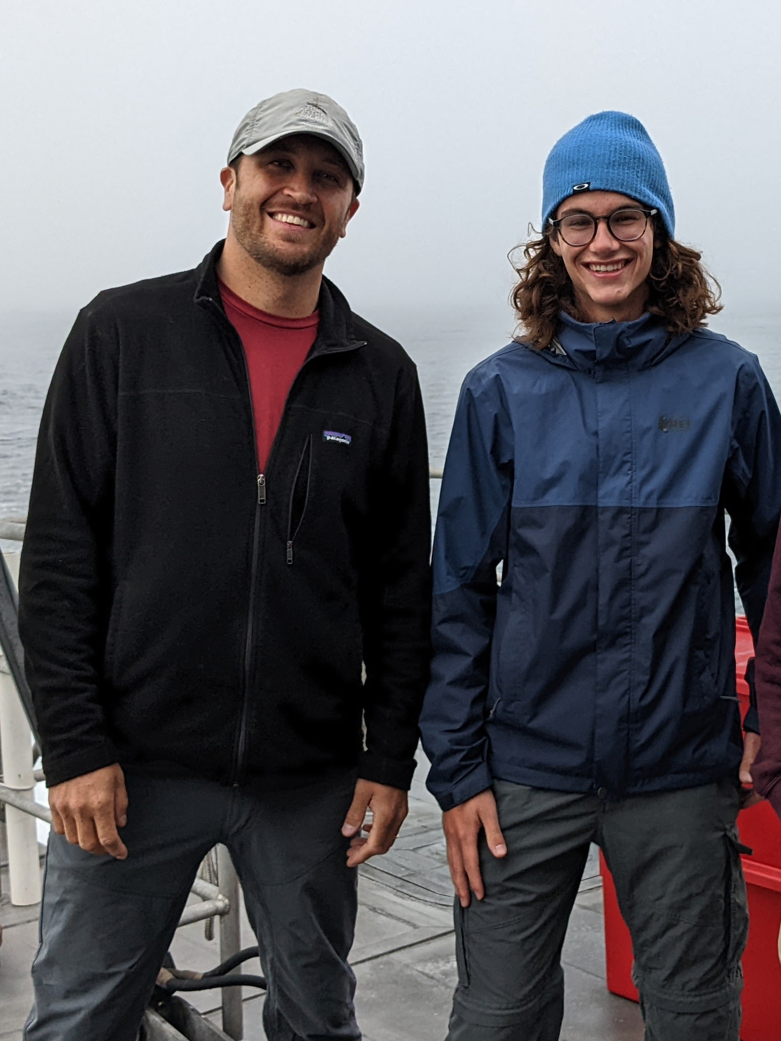 A professor and student smile for a photo together on a gray morning with the ocean behind them.