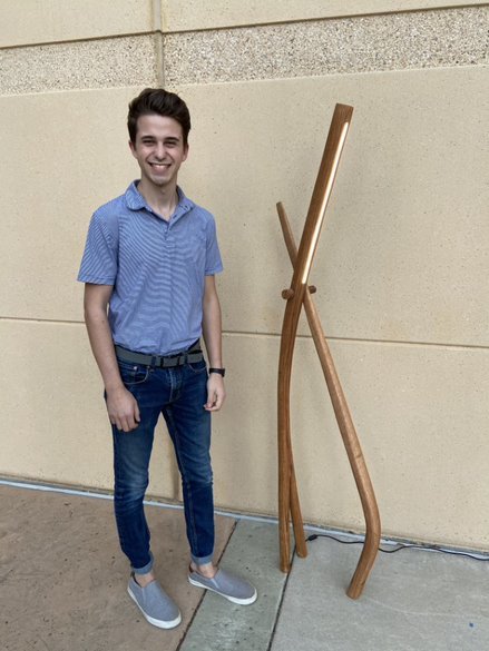 A male student stands next to his entry, a wooden floor lamp shaped like a wishbone.