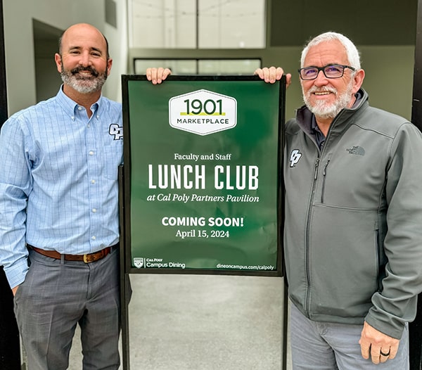 Two men stand by the Lunch Club sign