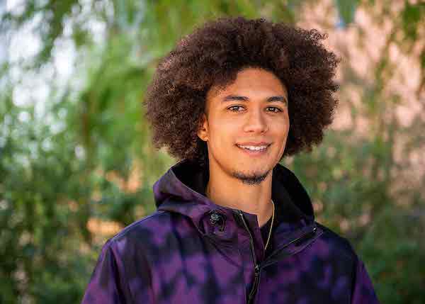A man with an afro and a goatee smiles while wearing a purple and black tie dye sweatshirt.