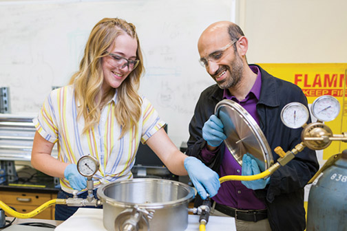  A student and a faculty member wearing eye protection handle a piece of metal equipment in an engineering lab