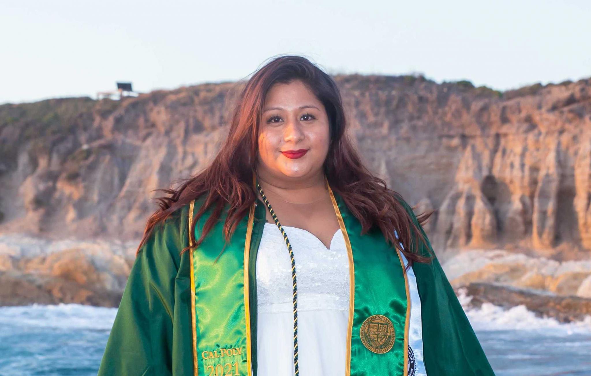 New graduate Francisca Camarillo stands in her green graduation robes in front of a beach with cliffs in the background.