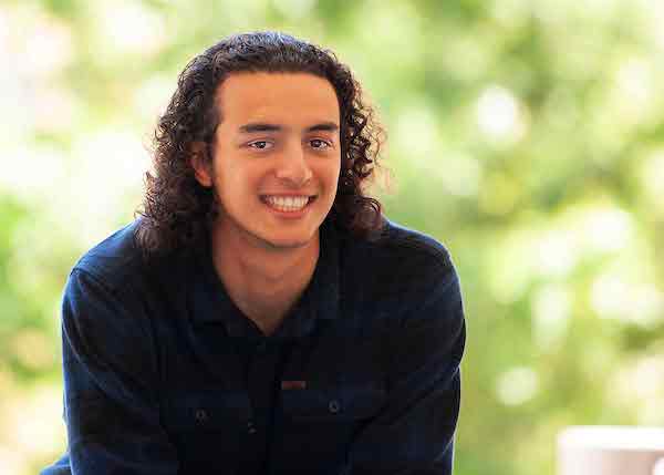 A man with slicked back curly hair smiles and wears a navy blue long sleeve shirt.