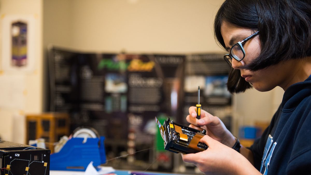 Student holds a screwdriver and works on a Cube Sat device.