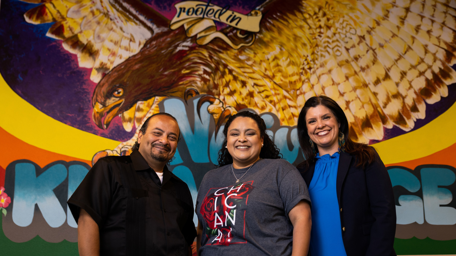 Cheryl Flores stands with Professors José and Jenell Navarro in front of a colorful mural in the Native American and Indigenous Cultural Center