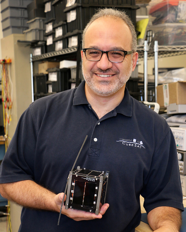 A man in a blue polo shirt smiles as he holds a 4-inch CubeSat in one hand.