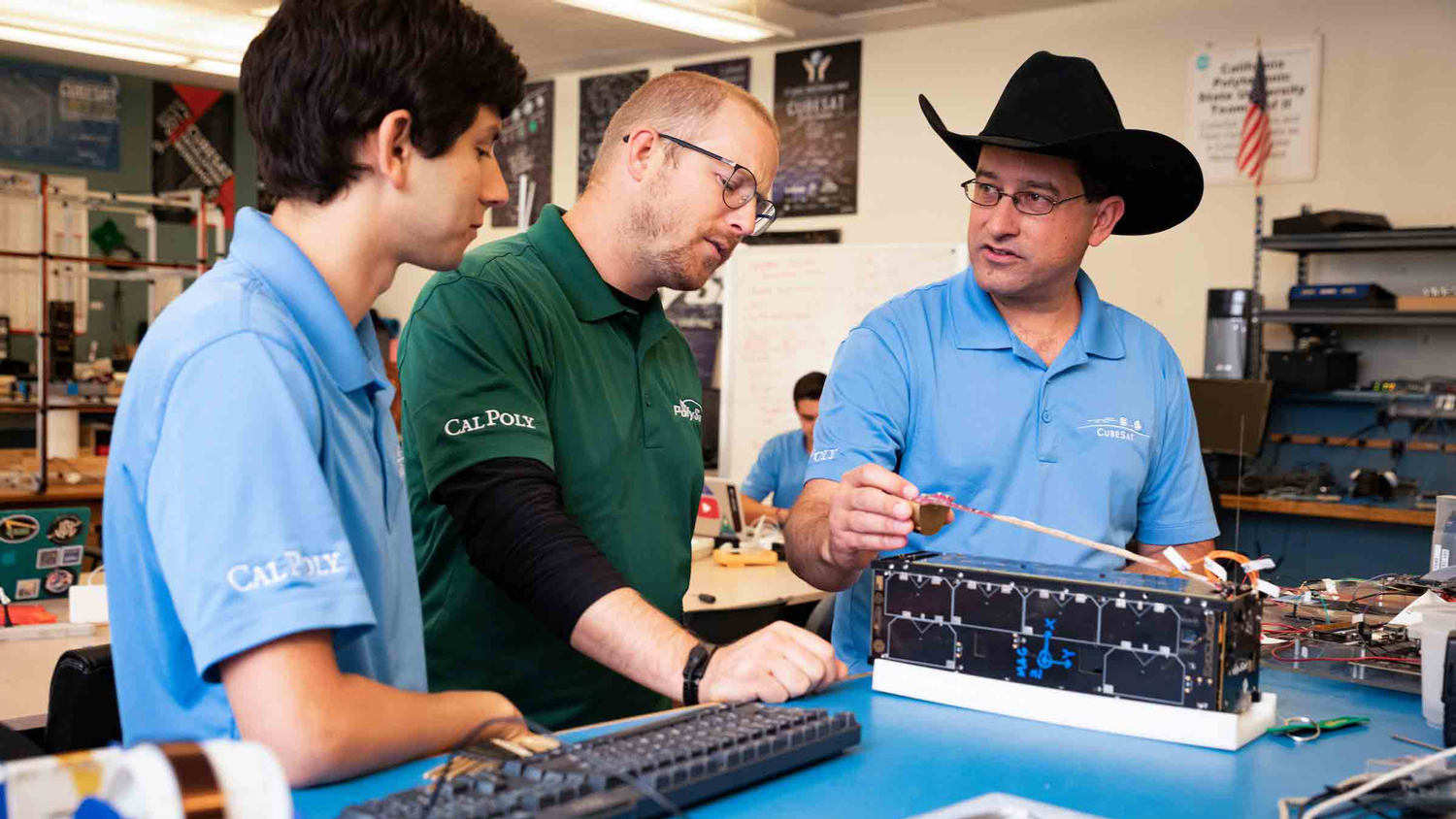 Three men, two in blue polo shirts and one in a green polo shirt, talk as they look at a CubeSat on the table in front of them.
