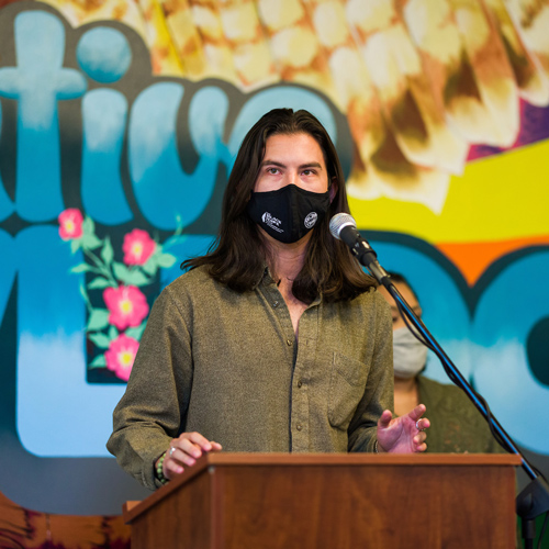 A man wearing a face covering stands in front of a colorful mural while he speaks