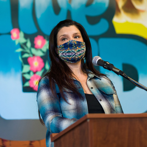 A person wearing a face mask speaks in front of a colorful mural. 