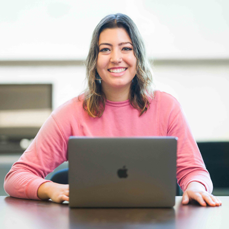 A person wearing a pink sweater sits in front of a laptop and smiles