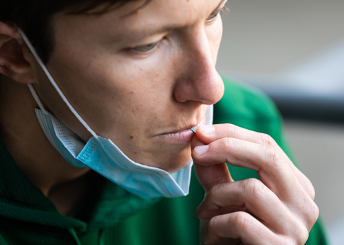 A student wearing a green sweatshirt inserts a saliva testing strip into his mouth