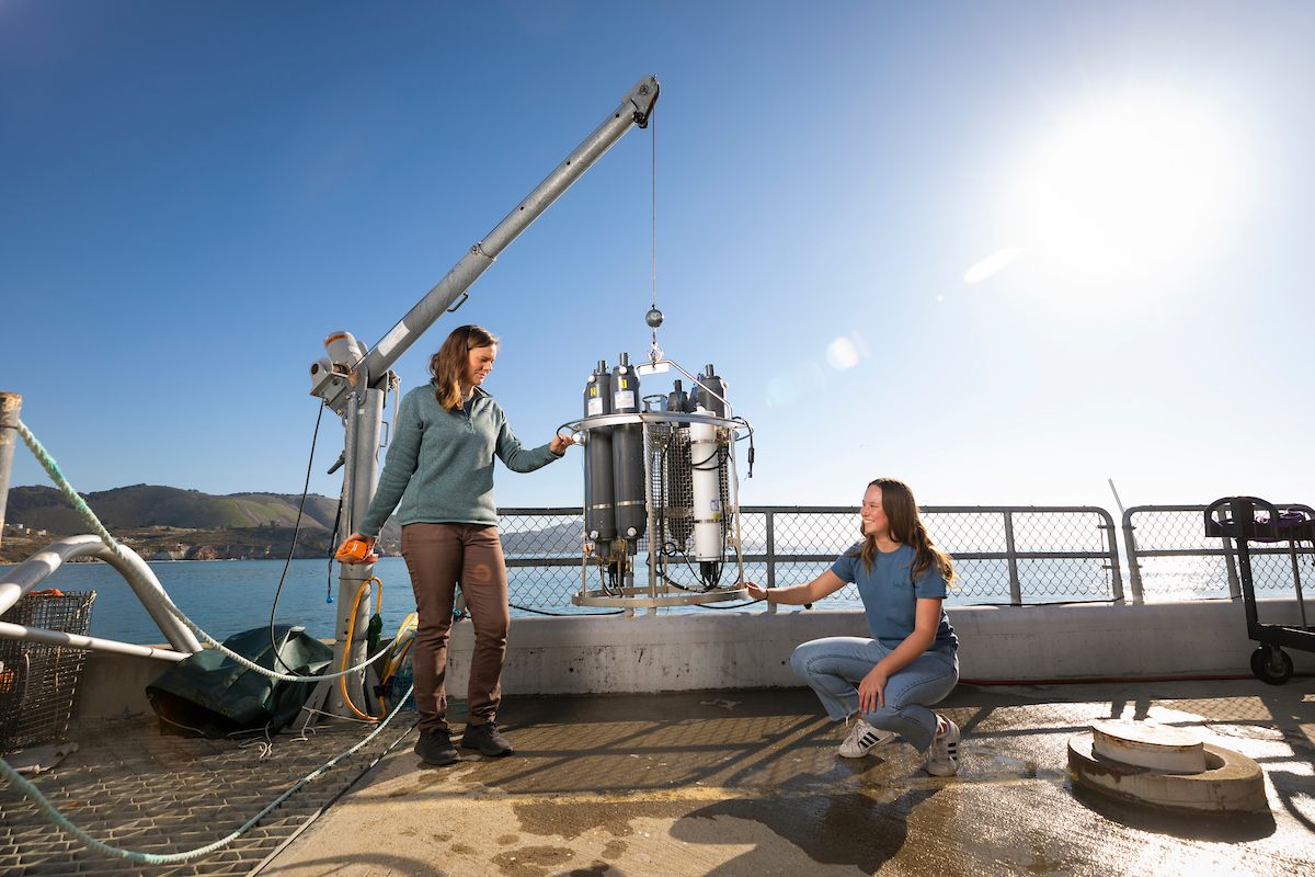 Two students stand on the Cal Poly Pier examining a tool that gathers water samples. 