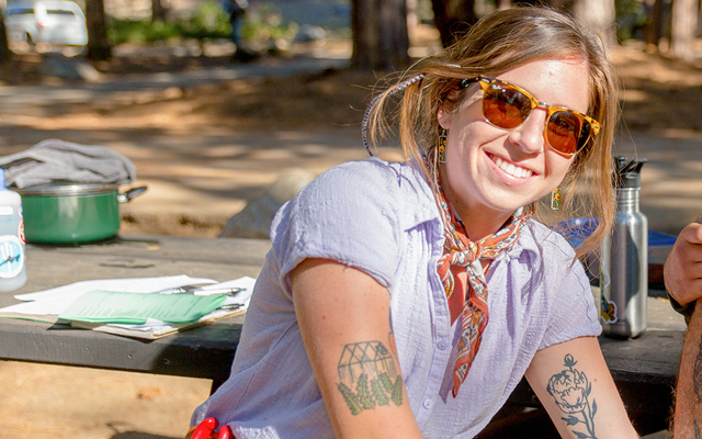 A woman in a gray shirt with plant-themed tattoos on both arms smiles at the camera.