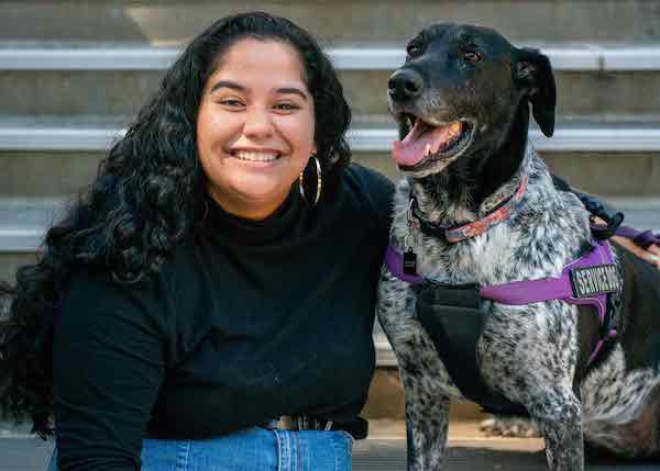 A woman with curly black hair wearing blue jeans, a black long sleeve turtleneck and hoop earrings sits and smiles next to a dog who is mostly black with some black and white patterning. The dog is wearing a purple and black vest that says 'Service Dog'