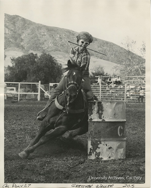A woman in old fashioned dress rides a horse around a barrel in a black-and-white archival photo.