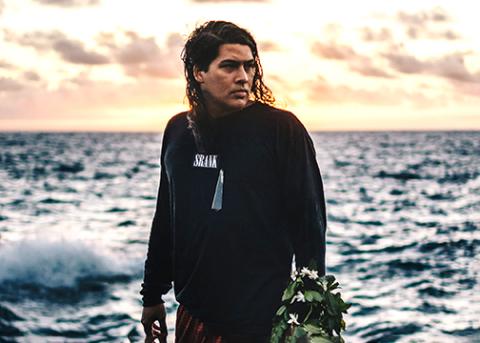 Poet William Nuutupu Giles stands at the edge of the ocean with a tropical sunset behind