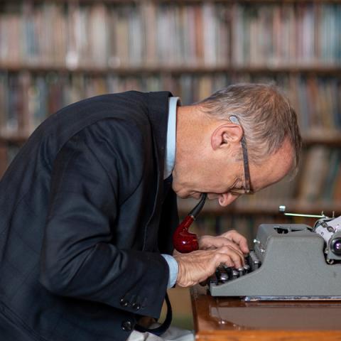 David Sedaris with glasses hunched over a typewriter with a pipe in his mouth typing on a typewriter
