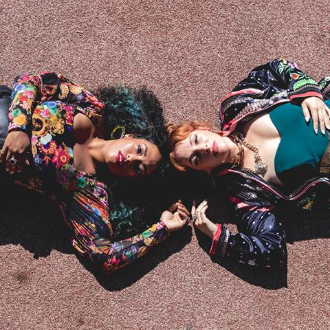 Photo of two female musicians laying on ground w the tops of their heads touching