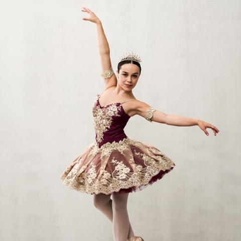 A female performer in a maroon ballet tutu holds her arms aloft in a dance pose