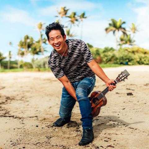 Jake Shimabururo plays ukulele on beach with palms in background
