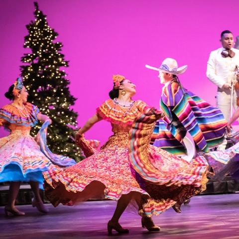 People on stage wearing traditional Mexican costumes dance in front of a lighted Xmas tree