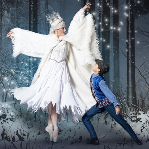 Woman in all-white dress and crown poses next to young boy in front of a snowy forest background