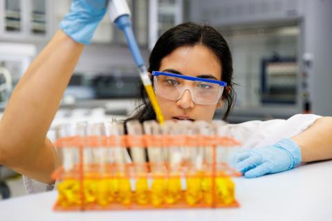 A young woman in safety glasses and latex gloves uses a lab syringe to add yellow liquid to a series of vials on a lab table
