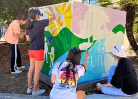 Four students paint a colorful mural on a large utility box on campus