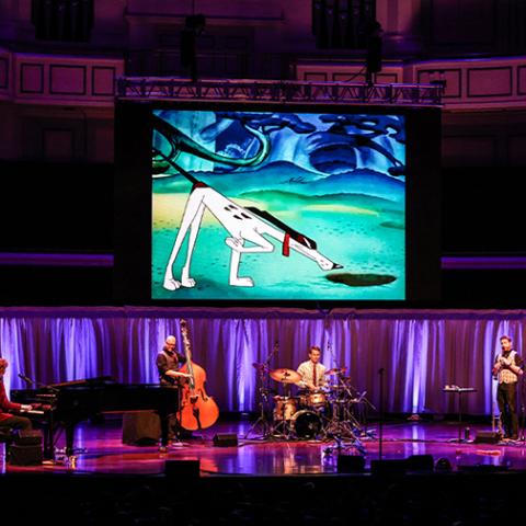 Band members play instruments on stage with a screen above showing a dog cartoon